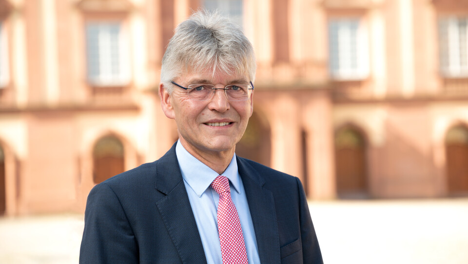 Prof. Spengel has short grey hair and is wearing a suit and tie. The castle can be seen in the background.