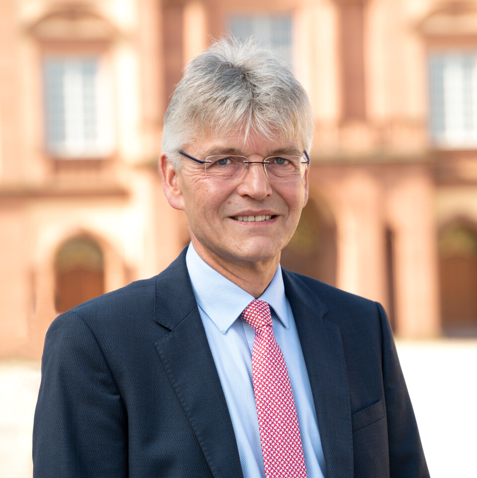 Prof. Spengel has short grey hair and is wearing a suit and tie. The castle can be seen in the background.