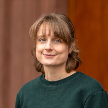 Sina Dreisbach hat schulterlange blonde Haare und trägt ein dunkelgrünes Oberteil