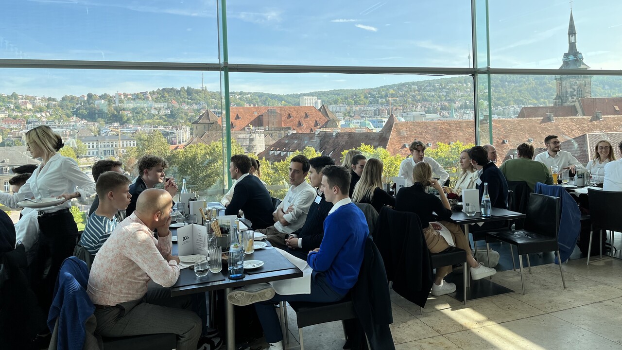 People are sitting at tables on the roof terrace of a restaurant. You can see rooftops through the glass wall.