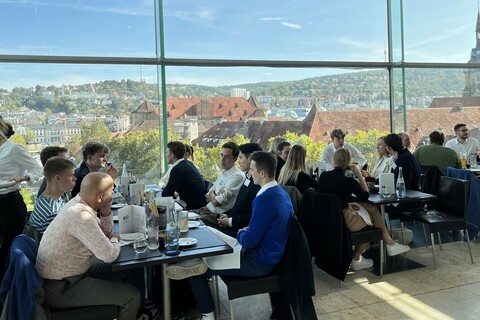 People are sitting at tables on the roof terrace of a restaurant. You can see rooftops through the glass wall.