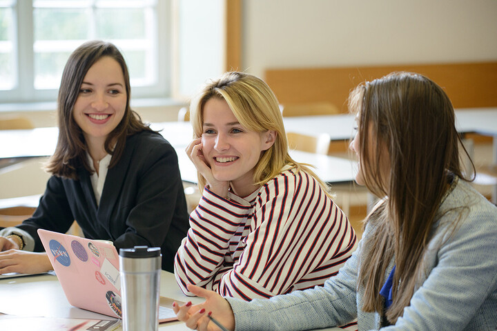 Drei Studentinnen sitzen lachend in einem Seminarraum.