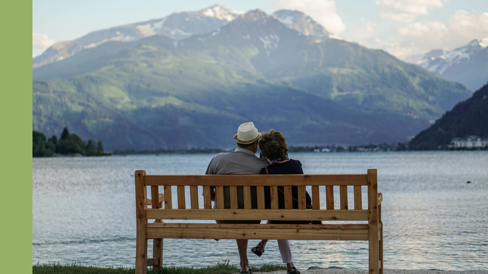 Zwei ältere Menschen sitzen Arm in Arm auf einer Bank. Sie blicken auf einen See und Berge