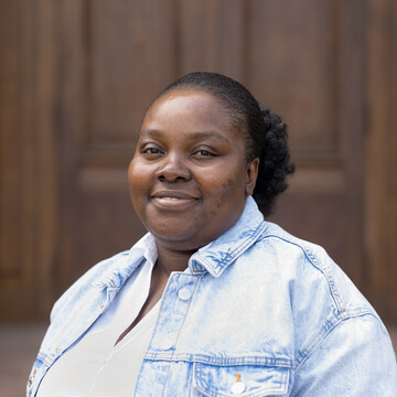Henrietta Oppong-Adjei. Sie trägt ein weißes Hemd und eine blaue Jacke und steht vor einer Holztür.