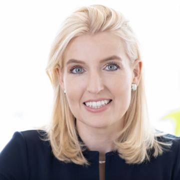 Claudia hat schulterlange blonde Haare, blaue Augen und trägt einen blauen Blazer