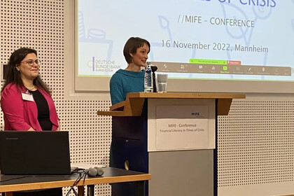 Carmela Aprea und Tabea Bucher-Koenen stehen an einem Podium vor einer Leinwand und dozieren.