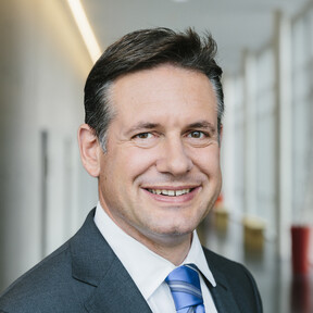Alexander Schwörer hat dunkelbraune, leicht graue Haare und braune Augen. Er lächelt und trägt ein weißes Hemd, ein dunkelgraues Sakko und eine blau, weiß gestreifte Kravatte.