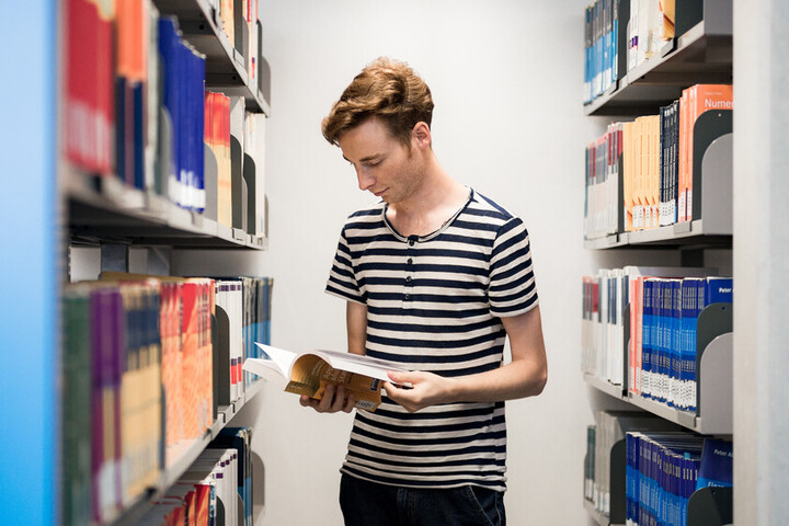 Student steht zwischen Bücherregalen und liest in einem Buch
