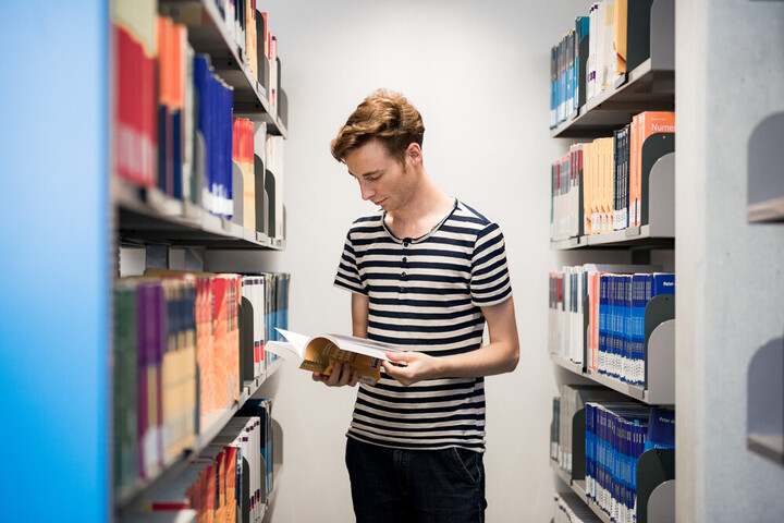 Student steht zwischen Bücherregalen und liest in einem Buch