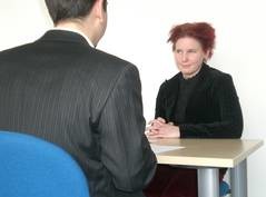 An einem Tisch sitzen sich eine Frau mit kurzen roten Haaren und schwarzem Anzug sowie ein Mann mit schwarz-grauen Haaren und antrazit-farbenem Anzug gegenüber.