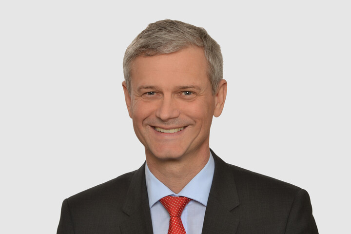 Prof. Helmig hat kurze graue Haare und trägt ein dunkles Jacket, ein hellblaues Hemd und eine rote Krawatte.