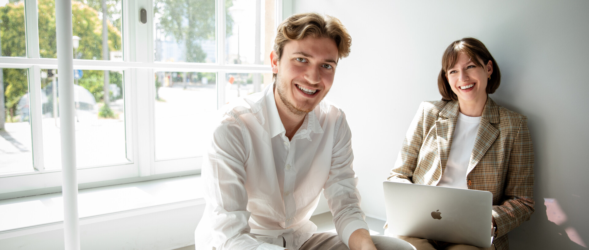 Turning talent into impact. 2 Studierende sitzen mit Laptop auf einer Fensterbank und werden von der Sonne angestrahlt