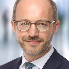 Herr Wagner hat graue Haare, eine Brille und trägt ein weißes Hemd mit schwarzem Anzug und blauer Krawatte