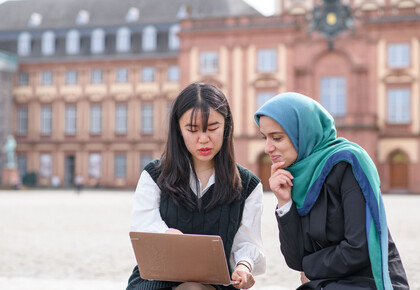 Zwei internationale Studentinnen sitzen auf dem Ehrenhof vor dem Schloss und blicken auf einen Laptopbildschirm