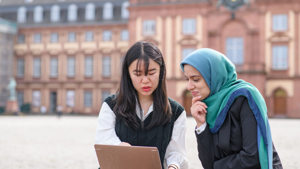 Zwei internationale Studentinnen sitzen auf dem Ehrenhof vor dem Schloss und blicken auf einen Laptopbildschirm