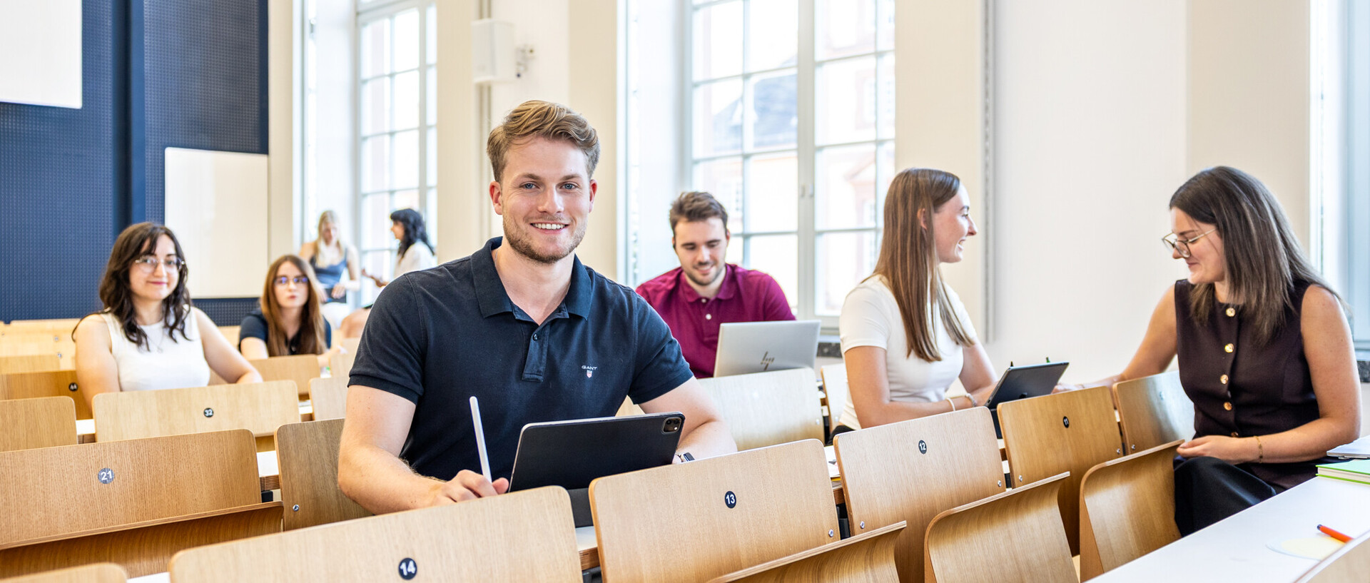 Turning talent into impact. Junger Mann mit blauem T-Shirt und blondem Haar sitzt in einem Hörsaal und schaut in die Kamera.