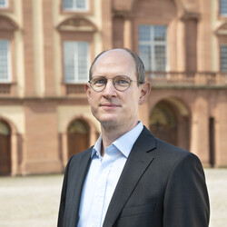 Prof. Dr. Oliver Spalt has short hair and glasses. He is wearing a light-colored shirt and a dark sacco and is standing in the Ehrenhof
