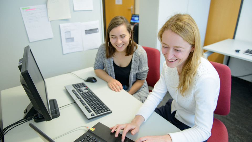 Zwei Studentinnen sitzen lachend an einem Tisch nebeneinander vor einem Laptop und einem Bildschirm mit einer externen Tastatur.