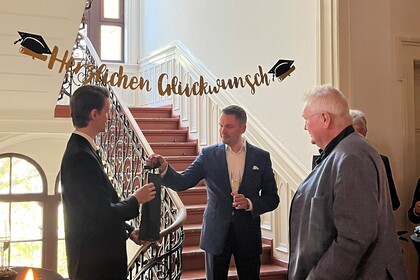Man sieht Sebastian Junker, Pro. Florian Kraus und Prof. Christian Homburg. Florian Kraus übergibt Herrn Junker eine Flasche Wein zur bestandenen Promotion
