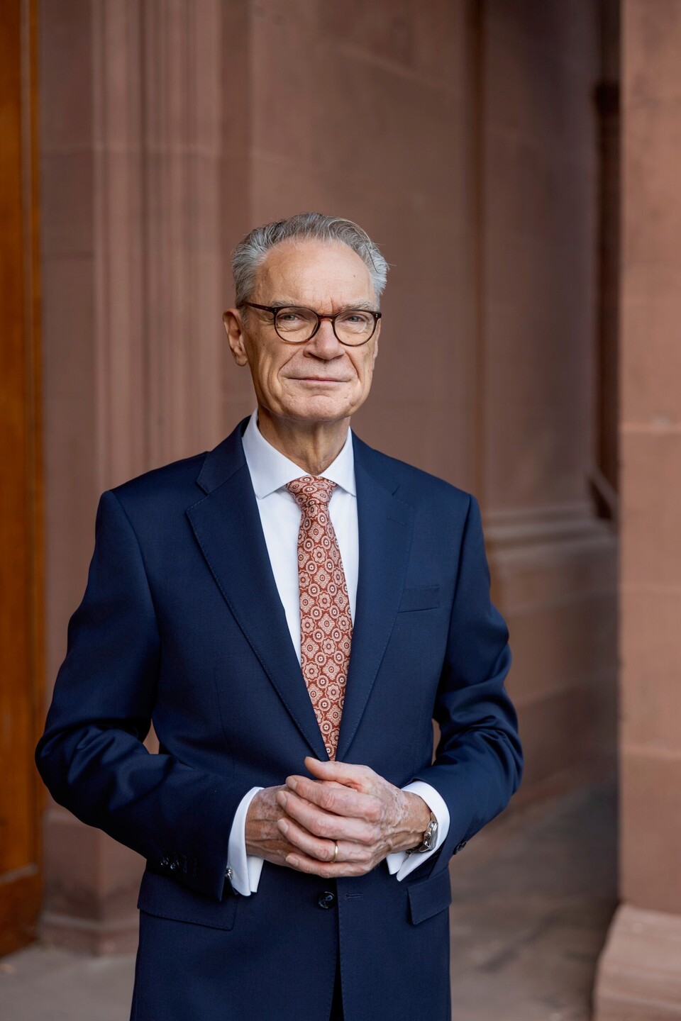 Joachim Lutz hat kurze Haare und trägt eine Brille und einen dunklen Anzug. Er steh mit verschränkten Händen vor einer Backsteinwand