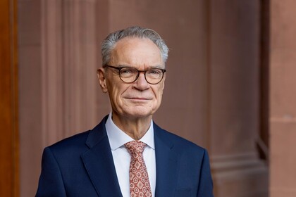 Joachim Lutz hat kurze Haare und trägt eine Brille und einen dunklen Anzug. Er steh mit verschränkten Händen vor einer Backsteinwand
