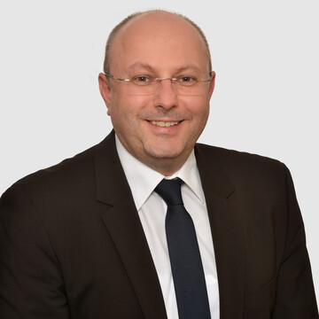 Prof. Stolletz hat eine Brille und trägt einen dunklen Anzug mit Krawatte und weißem Hemd.