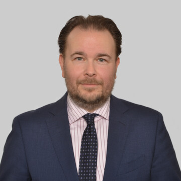 Herr Daske hat kurze braune Haare und trägt ein weißes gestreiftes Hemd mit blauem Anzug und Krawatte