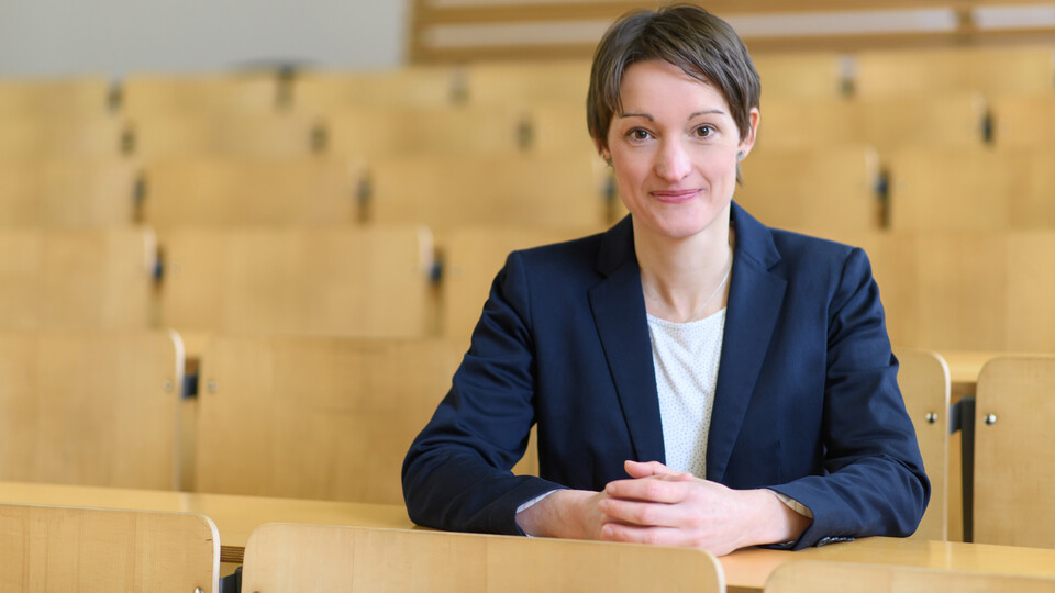 Portrait von Tabea Bucher-Koenen. Sie trägt eine weiße Bluse sowie einen dunkelblauen Blazer und sitzt mit gefaltetenen Händen in einem leeren Hörsaal.