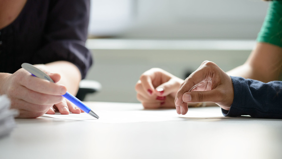 Zwei Studierende werden beraten, dabei sind die Hände und der Stift der Mitarbeiterin, der auf ein Papier zeigt, im Fokus.