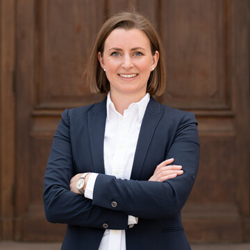 Portrait of Ms. Fischer. She is wearing a white blouse with a black blazer over it and is standing in front of a door of Mannheim Palace.
