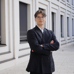 Finn Schütz trägt einen dunklen Anzug, hat hellbraune Haare und trägt eine Brille.