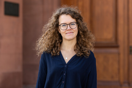 Frau Loetters hat braune lange gelockte Haarre eine Brille und trägt eine blaue Bluse