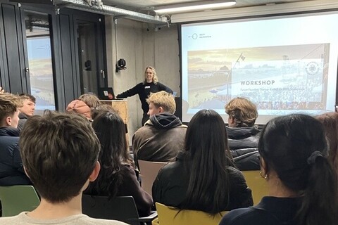 Eine Personengruppe von hinten. Die Menschen sitzen auf Stühlen und schauen auf eine Präsentation.