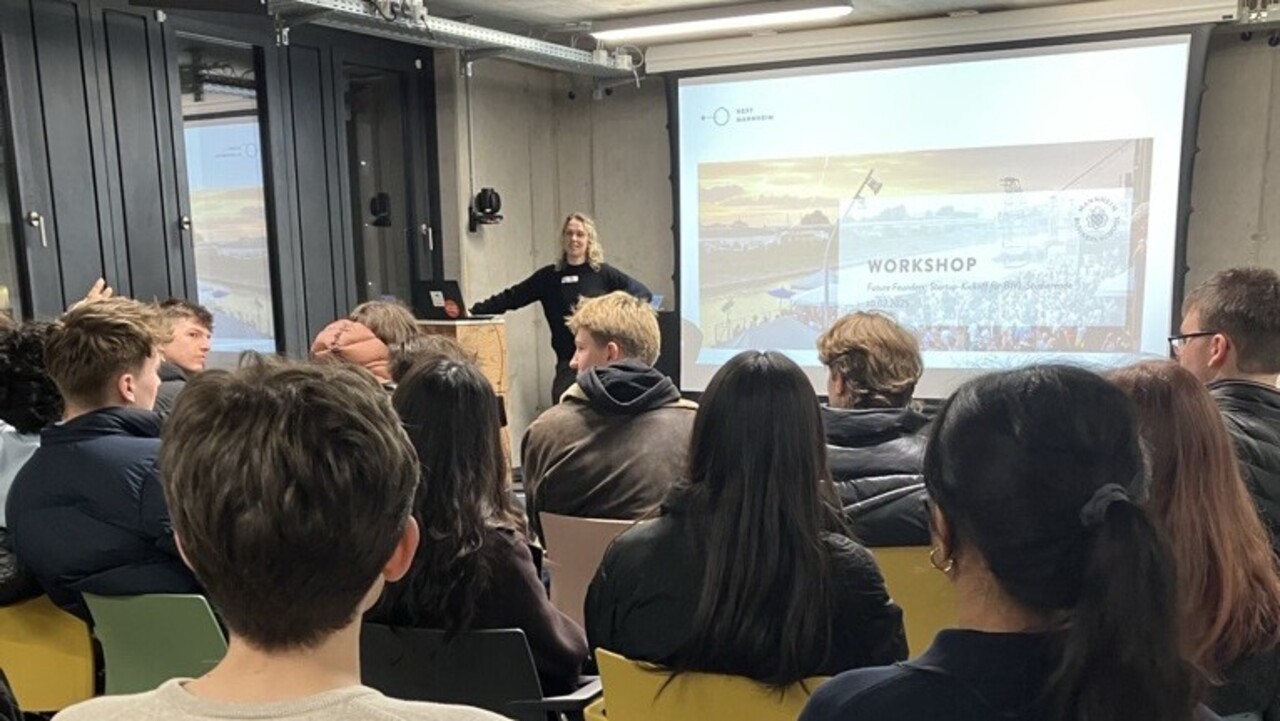 Eine Personengruppe von hinten. Die Menschen sitzen auf Stühlen und schauen auf eine Präsentation.