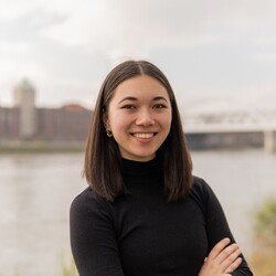 Paulina Wang has shoulder-length dark hair and is wearing a black turtleneck sweater. She has her arms crossed in front of her chest and is smiling. A river can be seen in the background