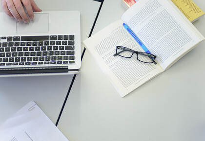 Auf einem aufgeschlagenen Buch liegt eine Brille und daneben steht ein Laptop.