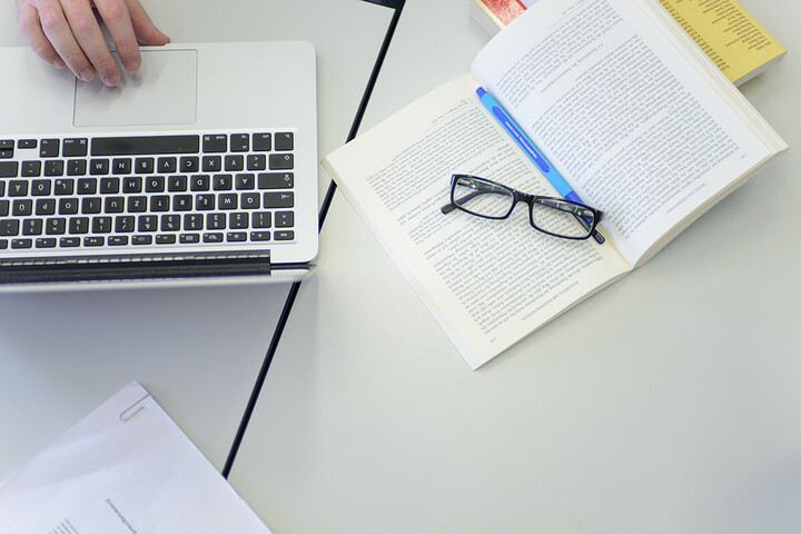 Auf einem aufgeschlagenen Buch liegt eine Brille und daneben steht ein Laptop.