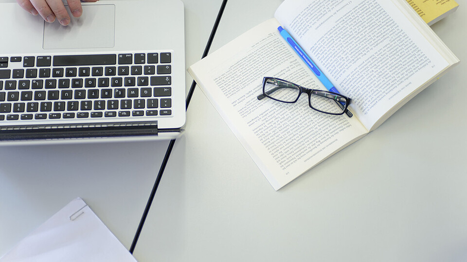 Auf einem aufgeschlagenen Buch liegt eine Brille und daneben steht ein Laptop.