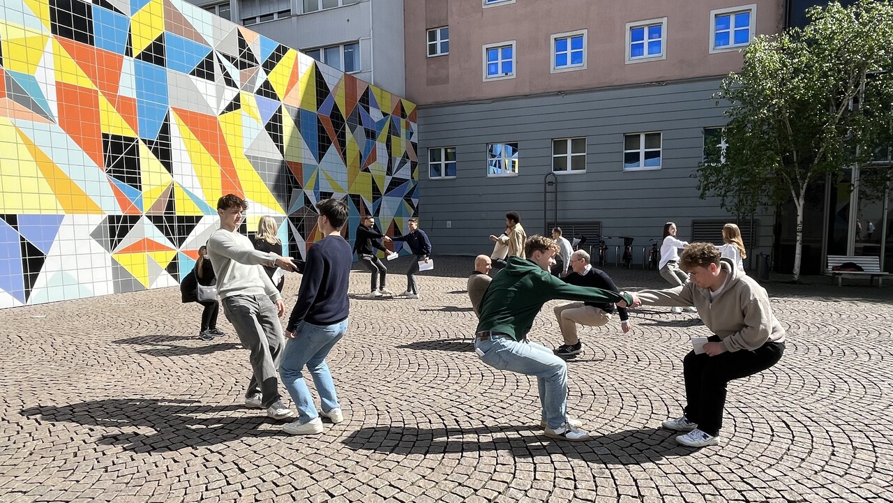 People in groups of two are holding hands and leaning backwards. In the background is a colorfully painted wall.