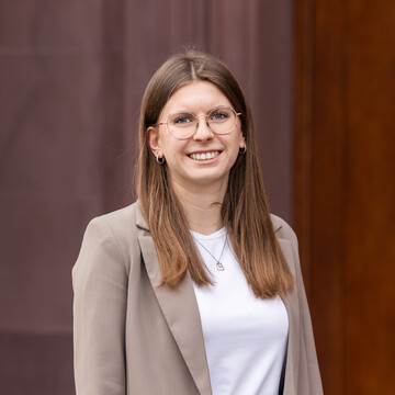 Sophie Peter has long brown hair and is wearing a white top and a beige blazer. She is standing in front of a dark red background and is smiling