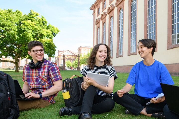 Drei Studierende sitzen auf der Mensawiese mit ihrem Laptop auf dem Schoß, mit dem Schloss im Hintergrund.