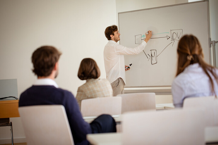 Eine Person steht am Whiteboard und erklärt drei anderen Personen, welche vor ihm sitzen den Zusammenhang zwischen unabhängiger, abhängiger und Mediatorvariable.