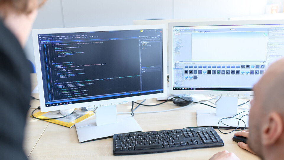 Zwei Forschende schauen auf zwei Monitore, auf denen Codes zu sehen sind.