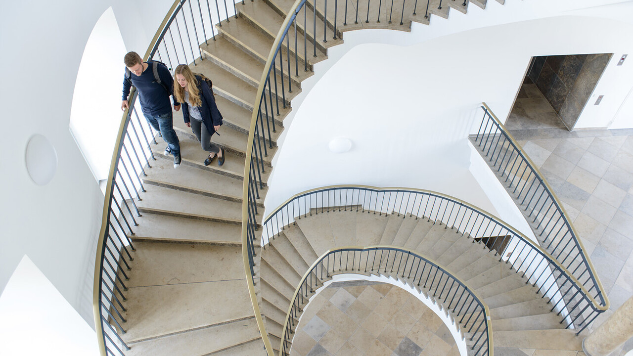Zwei Studierende laufen eine Wendeltreppe aus Stein in der Uni hinunter