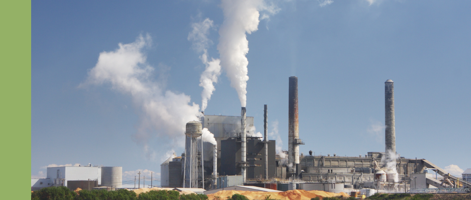Industrieanlage mit mehreren Schornsteinen, aus denen dichte weiße Rauch- bzw. Dampfwolken in den blauen Himmel aufsteigen; im Vordergrund sind niedrige Gebäude, Rohre und gelbliche Materialhaufen zu sehen.