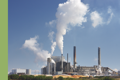Industrieanlage mit mehreren Schornsteinen, aus denen dichte weiße Rauch- bzw. Dampfwolken in den blauen Himmel aufsteigen; im Vordergrund sind niedrige Gebäude, Rohre und gelbliche Materialhaufen zu sehen.