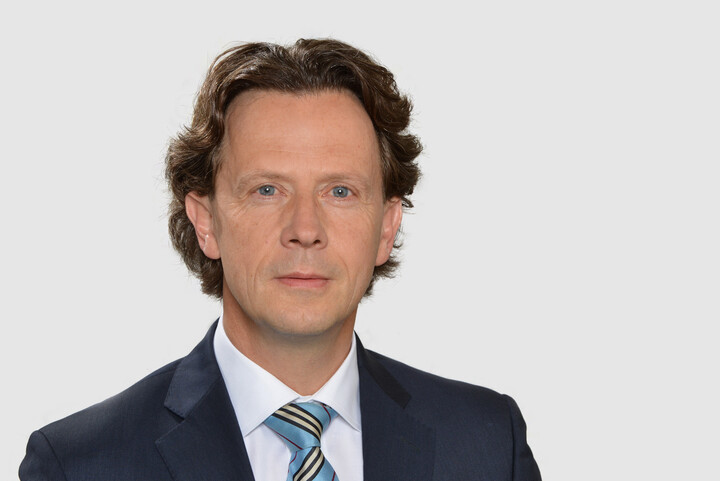 Michael Woywode hat leicht gewellte, etwas längere dunkle Haare. Er trägt ein helles Hemd, eine hellblaue Krawatte und ein blaues Jacket.