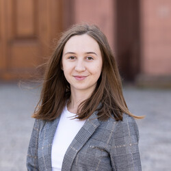 Dominika Galeziewska hat lange braune Haare und trägt eine weiße Bluse und einen grau karierten Blazer.