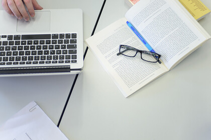 Auf einem aufgeschlagenen Buch liegt eine Brille und daneben steht ein Laptop.