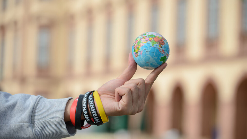 Eine Person hält einen Mini-Globus zwischen den Fingern, mit dem Schloss im Hintergrund.